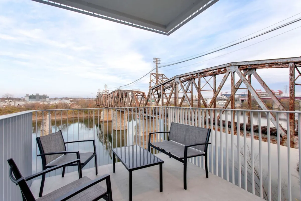 A peaceful balcony seating area with a clear view of the river and a rustic bridge. The space is inviting with chairs arranged for conversation or relaxation, and the golden hour light enhances the tranquil atmosphere. The setup hints at opportunities for small gatherings or personal reflection while enjoying the outdoors.