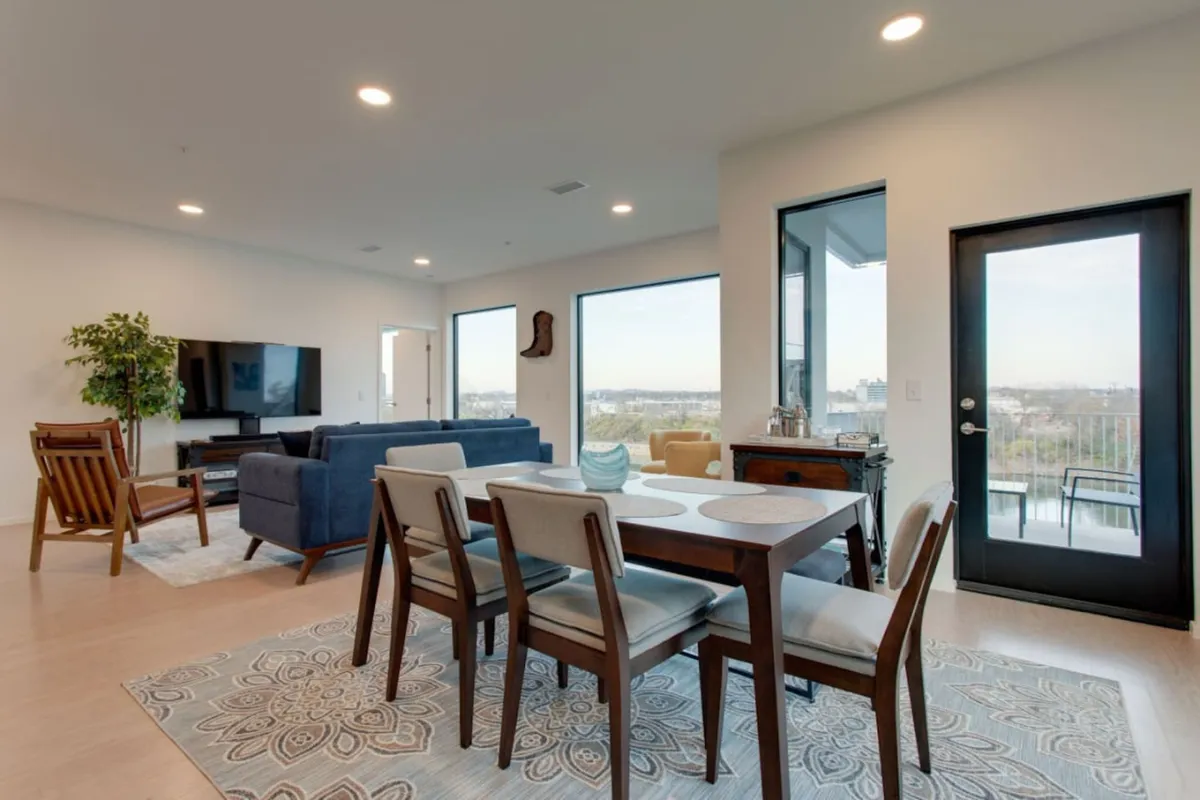 A spacious, sunlit open-plan living and dining area with a comfortable sofa, modern dining table, large windows offering city views, a television, and easy access to the balcony. The setup is ideal for groups to gather, relax, dine, and enjoy entertainment together.