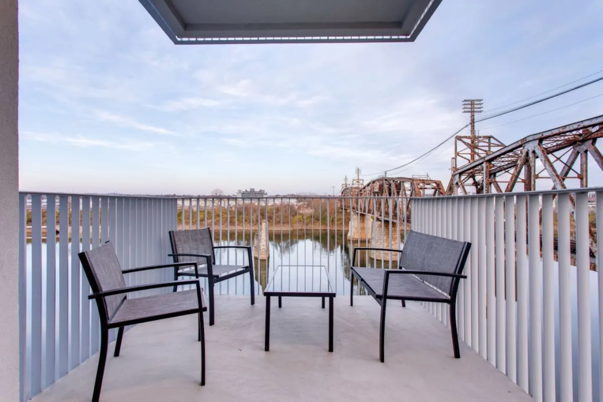 This image captures a balcony furnished with comfortable chairs and a table, overlooking a serene river with a prominent railroad bridge in the background. The scene is bathed in soft natural light, suggesting it could be either early morning or late afternoon, perfect for enjoying coffee, reading, or intimate gatherings. The urban skyline is visible in the distance, merging city energy with tranquil water views.