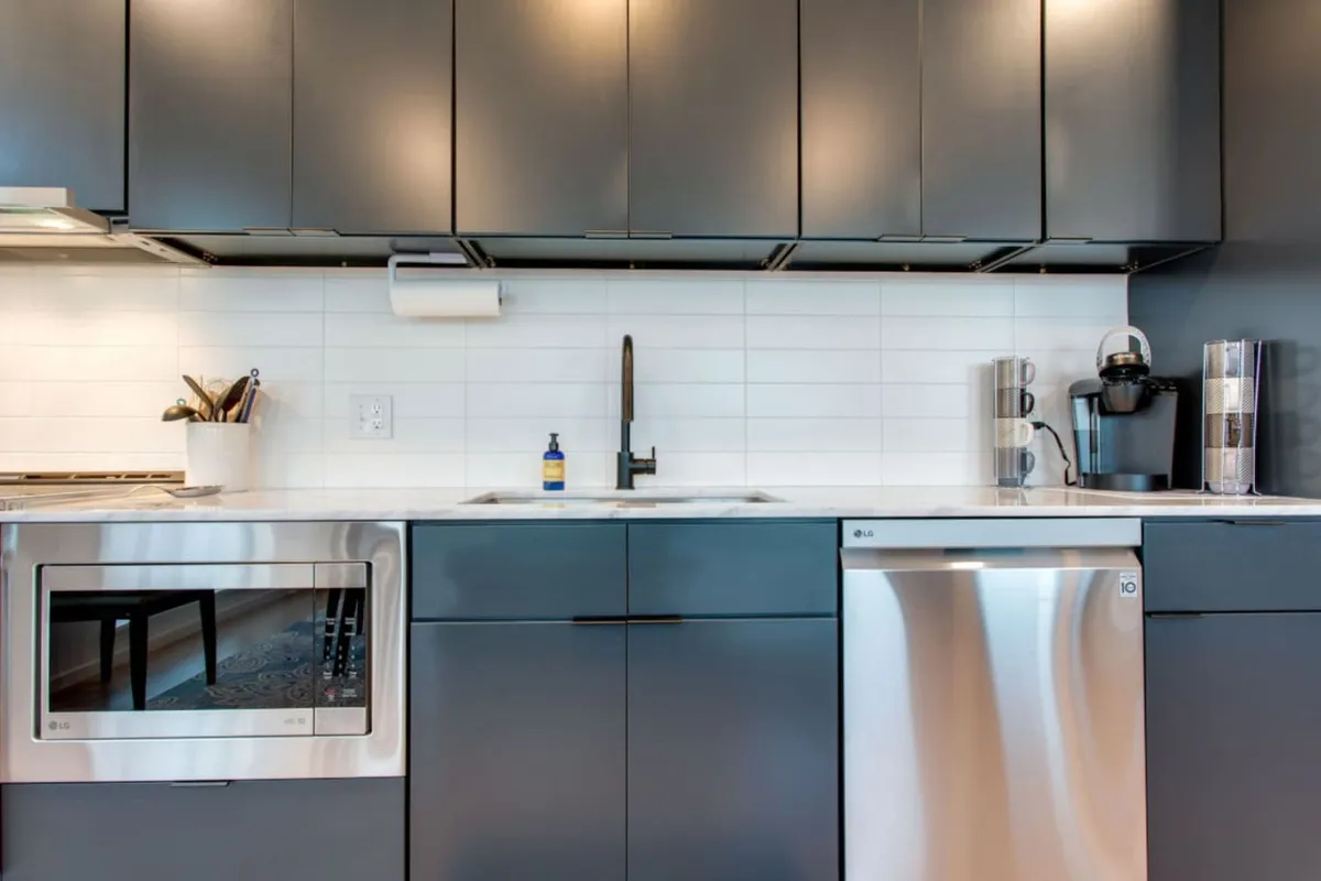 A close-up of the kitchen area focusing on stainless steel appliances, dark cabinetry, a bright white backsplash, and a visible coffee maker on the counter. The kitchen faucet and workspace emphasize both utility and comfort. The setup clearly highlights the dedicated coffee station and communal cooking possibilities.