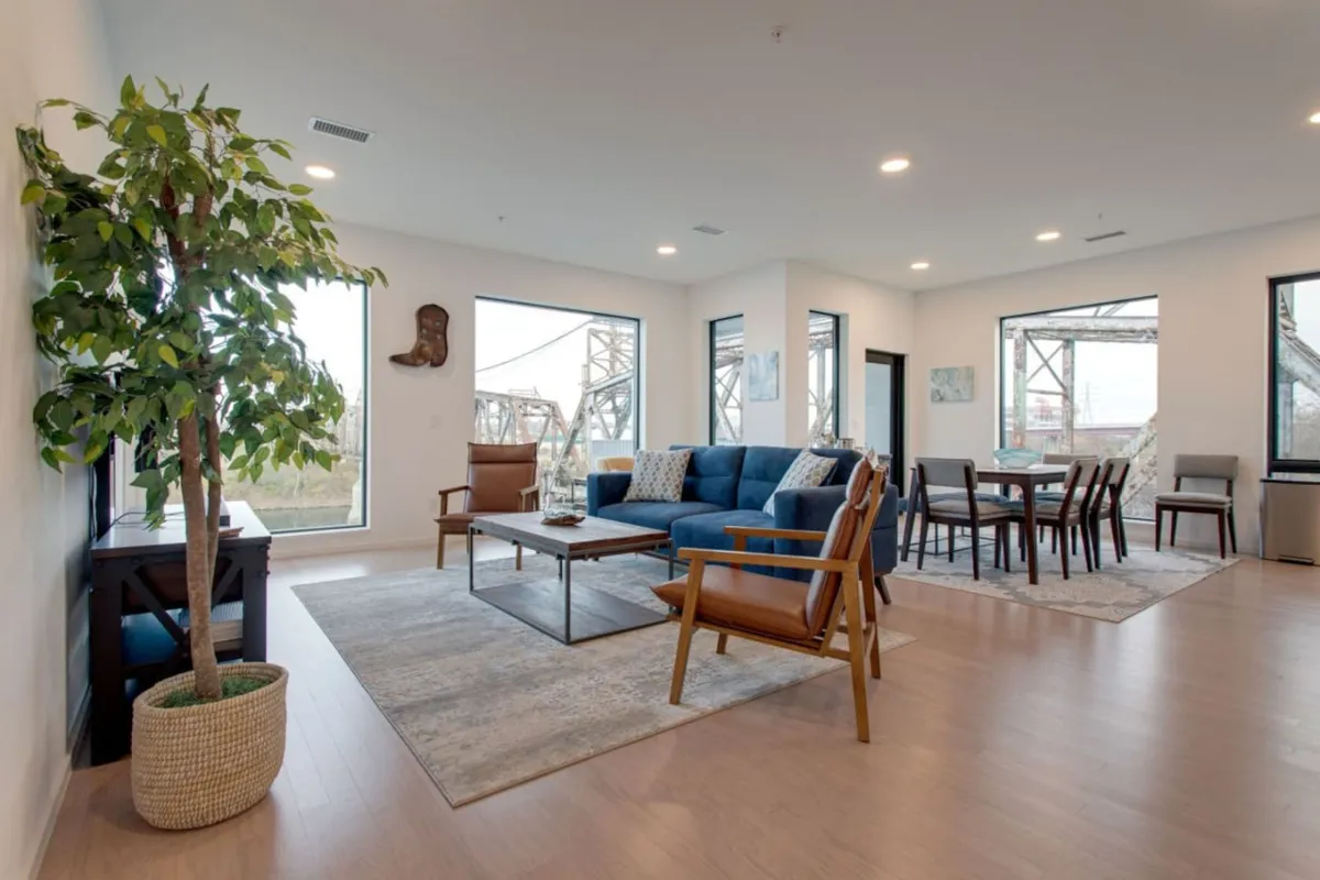A cozy living and dining space featuring a plush blue sofa, contemporary dining table, large windows, and greenery from an indoor plant. The room is inviting, with plenty of space for both social gatherings and quiet moments.