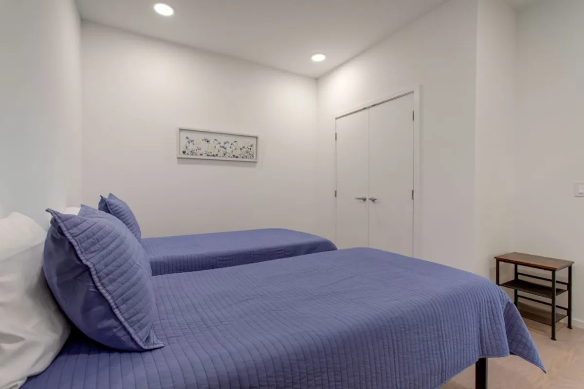 A cozy bedroom appointed with two twin beds dressed in blue quilts, complemented by a wooden bedside table and framed artwork. The space feels restful and cheerful, ideal for kids or singles seeking a quiet night.
