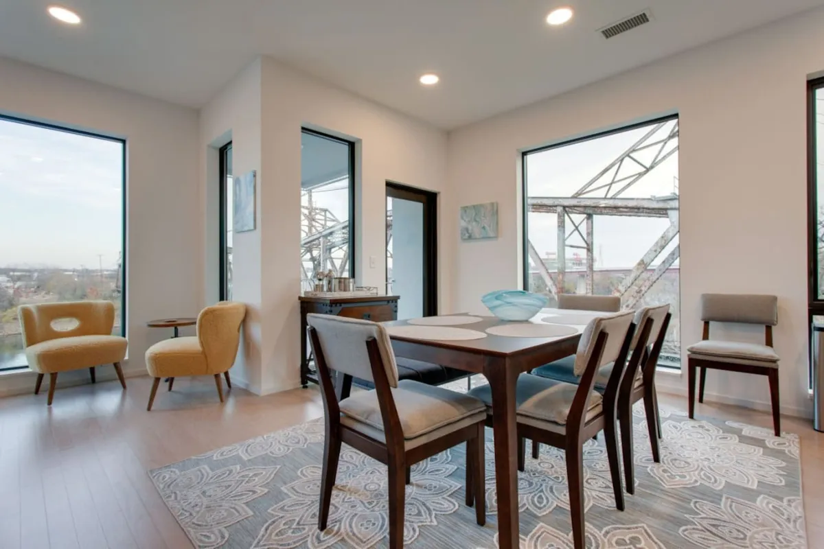 The image presents a contemporary dining table surrounded by chairs, situated near large windows with generous natural light and artistic touches. This setting is perfect for intimate gatherings or celebrations after exploring the city.