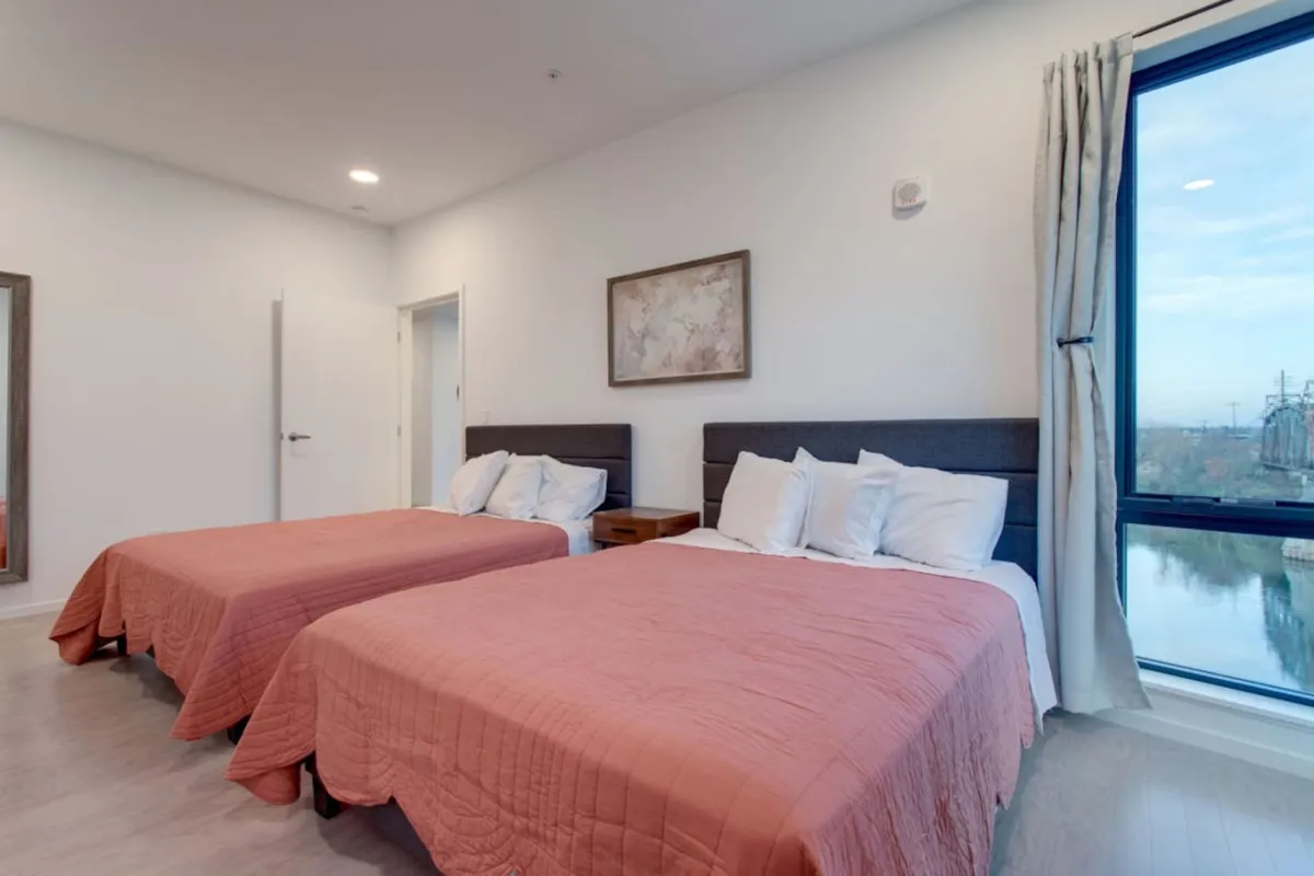 A welcoming bedroom with two beds featuring coral-colored bedding, a large window that bathes the room in sunlight, and artwork accentuating the contemporary feel. The space suggests ample comfort and a relaxing atmosphere ideal for families or groups of friends.