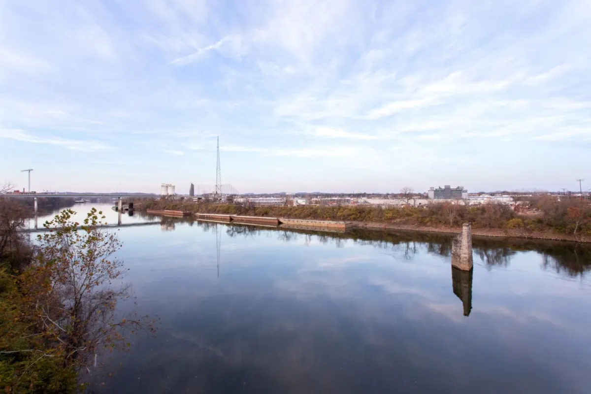 A tranquil riverfront scene with calm water, distant city buildings, bare trees, and telephone poles. The image evokes a sense of peace while nodding to the property's proximity to the river and city attractions.