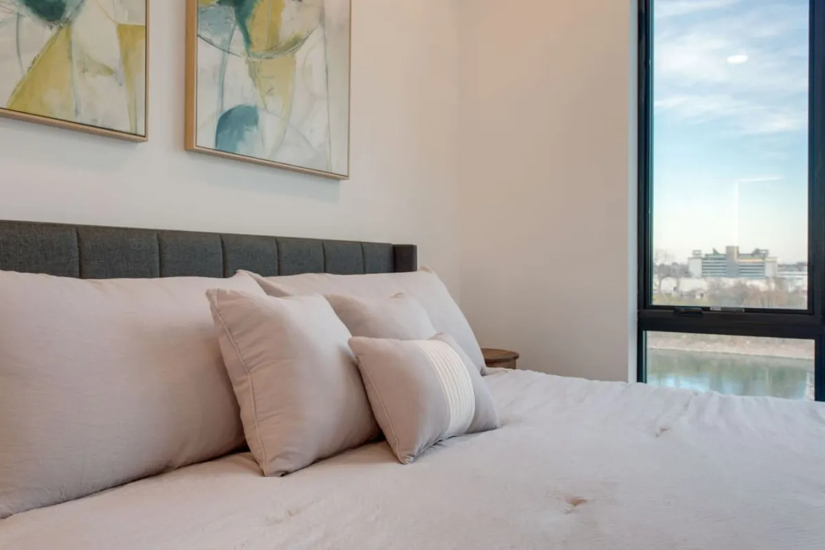 A tranquil bedroom scene with a comfortable bed adorned with decorative pillows, abstract art above the headboard, and a large window offering a view outside. The abundance of natural light and cozy atmosphere suggest a peaceful retreat amidst urban exploration.